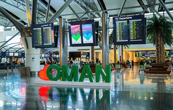 منظر داخلي لمطار مسقط الدولي مع لافتة "OMAN" وشاشات معلومات الرحلات