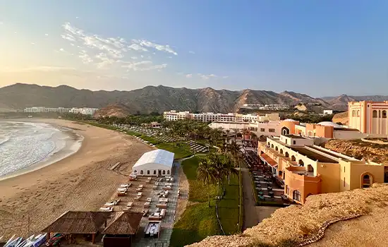 Exploring the Beaches of Oman: Which One Will You Enjoy the Most? 2 A view of Bandar Jissah Beach in Muscat, with golden sands, luxurious buildings, and towering mountains in the background.
