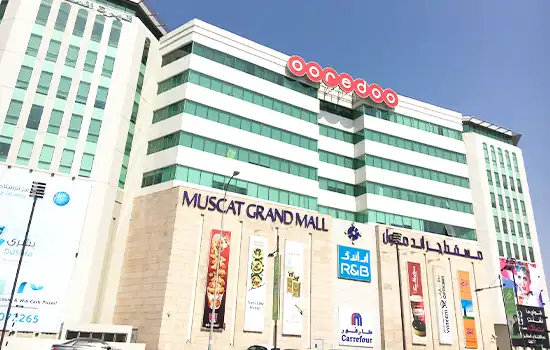 An exterior view of Muscat Grand Mall, showcasing modern architecture and popular retail brands, situated near neighboring shopping centers like Oman Mall and Avenues Mall.