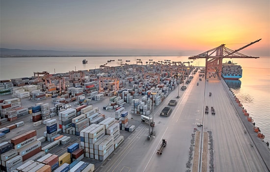 رتبه یازدهم اقتصاد جهانی برای شورای همکاری خلیج فارس؛ ۲.۱ تریلیون دلار تولید ناخالص و ۷۴۸ میلیارد دلار ذخایر ارزی 7 Aerial view of Port of Salalah during sunset, showing rows of shipping containers, cranes, and docked cargo ships.